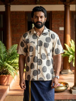 A man with a beard stands indoors, wearing the Dot Breeze Shirt—a beige half-sleeve shirt featuring oversized dot patterns, crafted from breathable lightweight fabric—and paired with a traditional blue sarong. Green potted plants and a bowl with flowers are in the background.