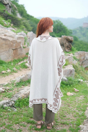 Back view of the model in the natural cream knit kimono, showing the plain knit back, the brown block printed collar and sleeve borders and the block printed panel running down the hem, over olive harem pants on a hillside path.
