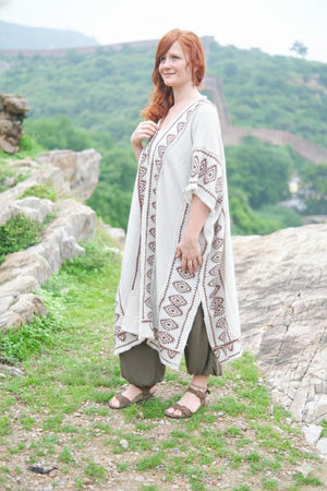 Full-length front view of the model in the natural cream knit kimono with the brown block printed border and cobweb fringe hem, worn open over olive harem pants with sandals, on a hillside path with green mountains behind.