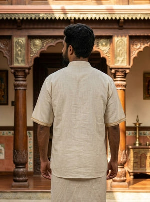 A man wearing the Midnight Nomad kurta—a beige short-sleeved traditional outfit featuring subtle handblock prints—stands with his back to the camera in front of an ornate wooden structure adorned with columns and intricate carvings.
