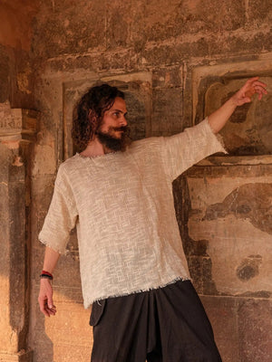 Beige cotton-jute baggy T-shirt on a model under a stone arch, raw frayed cuffs, one arm raised, dark trousers