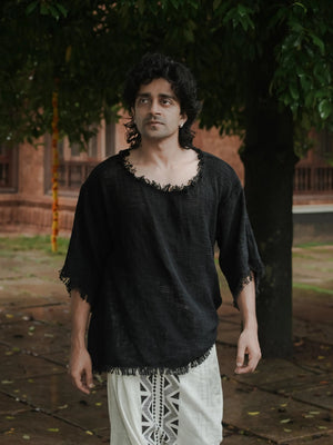 Black cotton-jute baggy knit T-shirt on a model walking under trees, frayed neck and sleeve hems, pale trousers