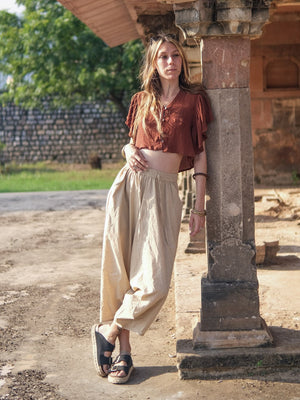 Beige cotton harem pants worn with a rust blouse and sandals, leaning on a carved sandstone pillar in warm daylight.