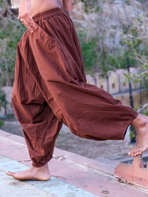 Rust brown harem pants stepping barefoot across a stone platform, gathered volume held above a narrow tapered ankle.