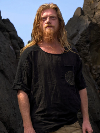 A man with long blond hair and a beard stands outdoors among dark rocks, wearing a loose Mandala T-shirt and light pants. The blue sky adds to his relaxed boho style.
