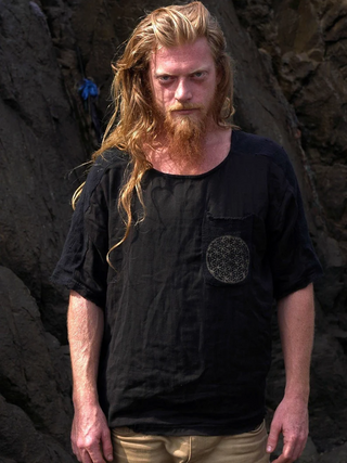A man with long, wavy red hair and a beard stands outdoors by rocky terrain, wearing a Mandala T-shirt with a round patterned pocket and light pants, gazing intently at the camera with a boho vibe.