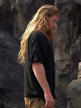 A person with long, wavy blond hair and a beard stands in profile against rocky mountains, wearing a sustainable Mandala T-shirt for a premium boho look and tan pants.