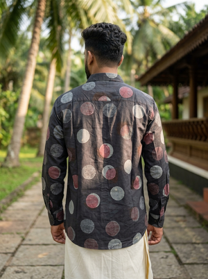 A man stands outdoors facing away, wearing the Aarush Polka Block Print Shirt—black with colorful polka dots—and a white traditional garment, seamlessly blending into a Boho wardrobe scene with palm trees and a wooden building in the background.