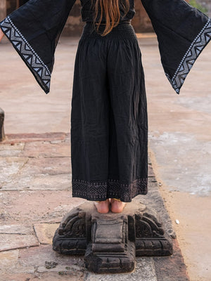 Model from behind in black Medusa palazzo pants and a black printed bell sleeve top, standing on a carved stone pedestal