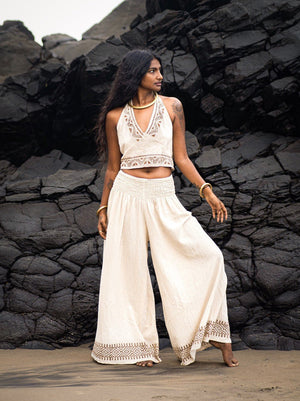 Model in natural Medusa palazzo pants and a cream embroidered halter crop top, standing before dark volcanic rocks