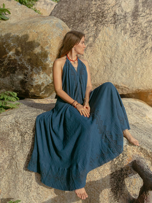 A woman in the Hanvi maxi dress, a long sleeveless blue gown made from sustainable cotton, sits barefoot on a large rock and gazes to the side. Sunlight illuminates her and the textured rocky backdrop.