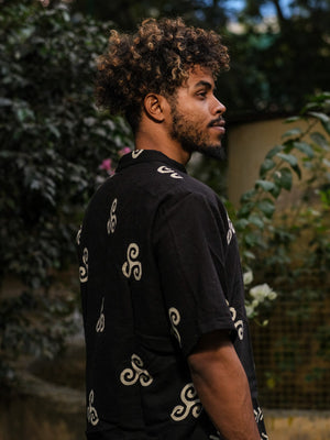 A man with curly hair and a beard stands in profile wearing a Spiral Kurta, surrounded by greenery in a dimly lit outdoor setting.
