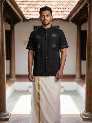 A man stands in a traditional courtyard, dressed in the Samhita Shirt—a black short-sleeve shirt adorned with distinctive block discharge prints—paired with a white dhoti featuring a yellow stripe. He faces forward, with wooden pillars and a tiled roof visible behind him.