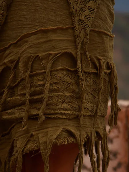 Close-up of the Titania Wrap Skirt in Olive Green, highlighted outdoors. This distinctive skirt showcases textured, layered fabric with rough edges, lace and fringe details, and hanging tassels.