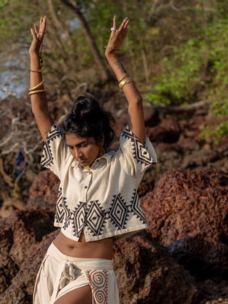 In a sunlit outdoor setting, a person with long hair raises their arms, wearing HWCs beige Tribal Crop Shirt featuring geometric black patterns. The scene of rocks and greenery perfectly complements the natural and stylish look.