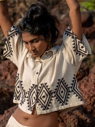A person wearing HWCs cream Tribal Crop Shirt featuring black geometric patterns stands with arms raised. The background is rocky and out of focus, and their dark hair is styled in an updo.