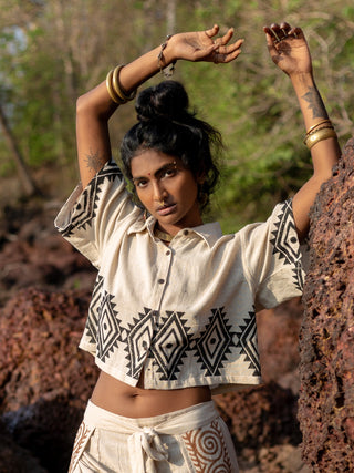 Wearing HWCs Tribal Crop Shirt, a person stands among rocks in the great outdoors. The beige outfit features black geometric tribal patterns, with their arms raised and bracelets adorning one wrist against a blurred greenery backdrop.