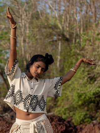 A person wearing an HWC Tribal Crop Shirt and matching pants joyfully dances outdoors with arms raised, surrounded by trees. Their bracelets and hair styled in a bun beautifully complement the tribal block design of their outfit.