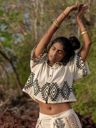 A person stretches outdoors in an HWC Tribal Crop Shirt and matching pants with geometric designs, surrounded by trees and greenery. Gold bangles on their wrists add elegance to the scene.