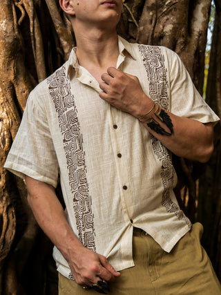Wearing the beige Utopian shirt with a tribal print, this person stands before large tree roots, holding the collar. The look is styled with light tan pants plus wrist and finger accessories.