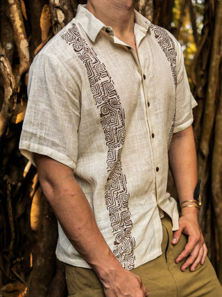 A person in a beige short-sleeved linen Utopian shirt with geometric brown tribal-inspired patterns, buttons, and a collar stands before tree roots, paired with light brown pants. The face is not visible.
