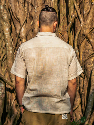 A person with short dark hair in a topknot wears a beige Utopian shirt and olive-green pants, standing with their back to the camera in front of large tree roots.