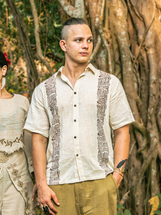 A person with light skin and a topknot stands outdoors in front of large tree roots, wearing a short-sleeved beige Utopian shirt with tribal-inspired patterned panels and tan pants. Another persons shoulder is partially visible beside them.