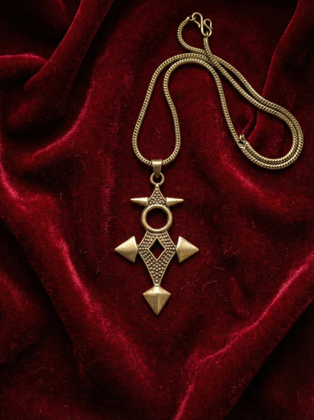 The Scroll Pendant makes a striking geometric statement jewelry piece, its bronze form featuring intricate shapes and resting elegantly on deep red velvet fabric.