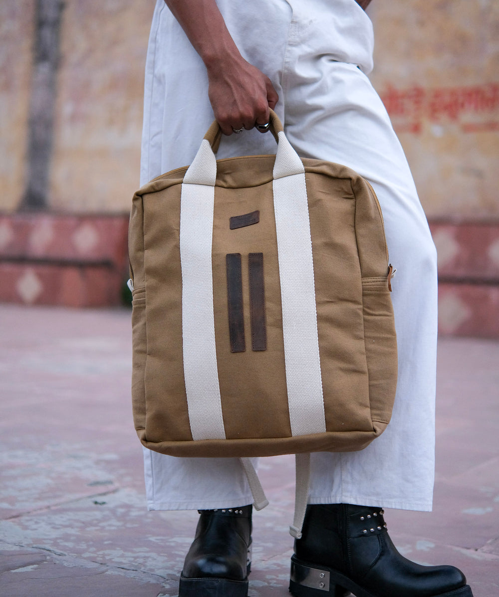 A person in white pants and black boots holds a Camel Brown Kelly Back Pack with two vertical white stripes while standing outdoors on a tiled surface.