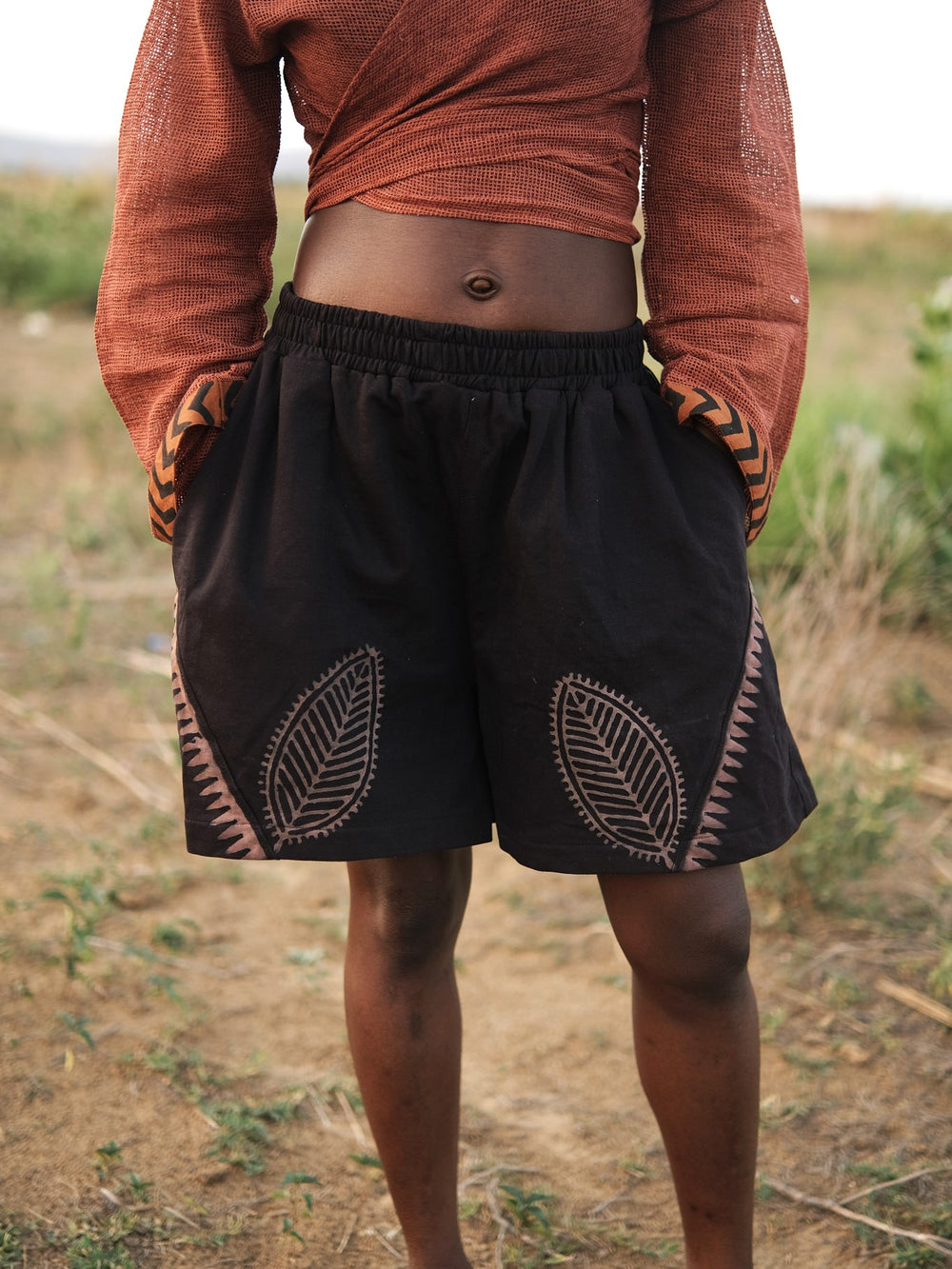 A person standing outdoors wears a long-sleeve rust-colored top paired with the Kaari Leaf Shorts, which showcase bold black leaf patterns. The grassy background is blurred, emphasizing the Kaari Leaf Shorts and outfit details from waist to knee.