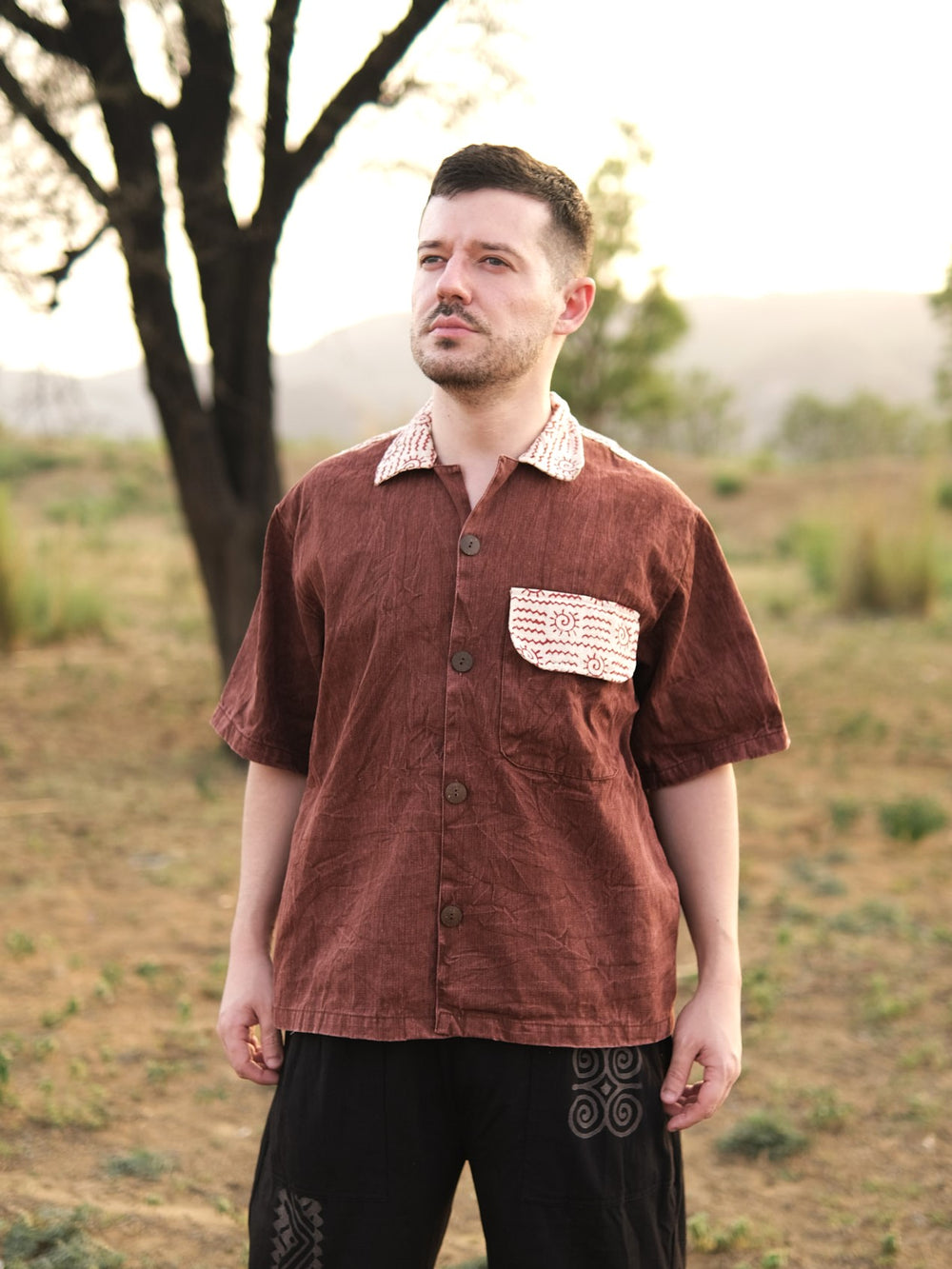A person stands outdoors on dry grass, wearing the Rustwood Shirt in coffee brown—a relaxed fit cotton shirt with patterned white accents on the collar and pocket—and dark pants. Trees and hills are visible in the background.