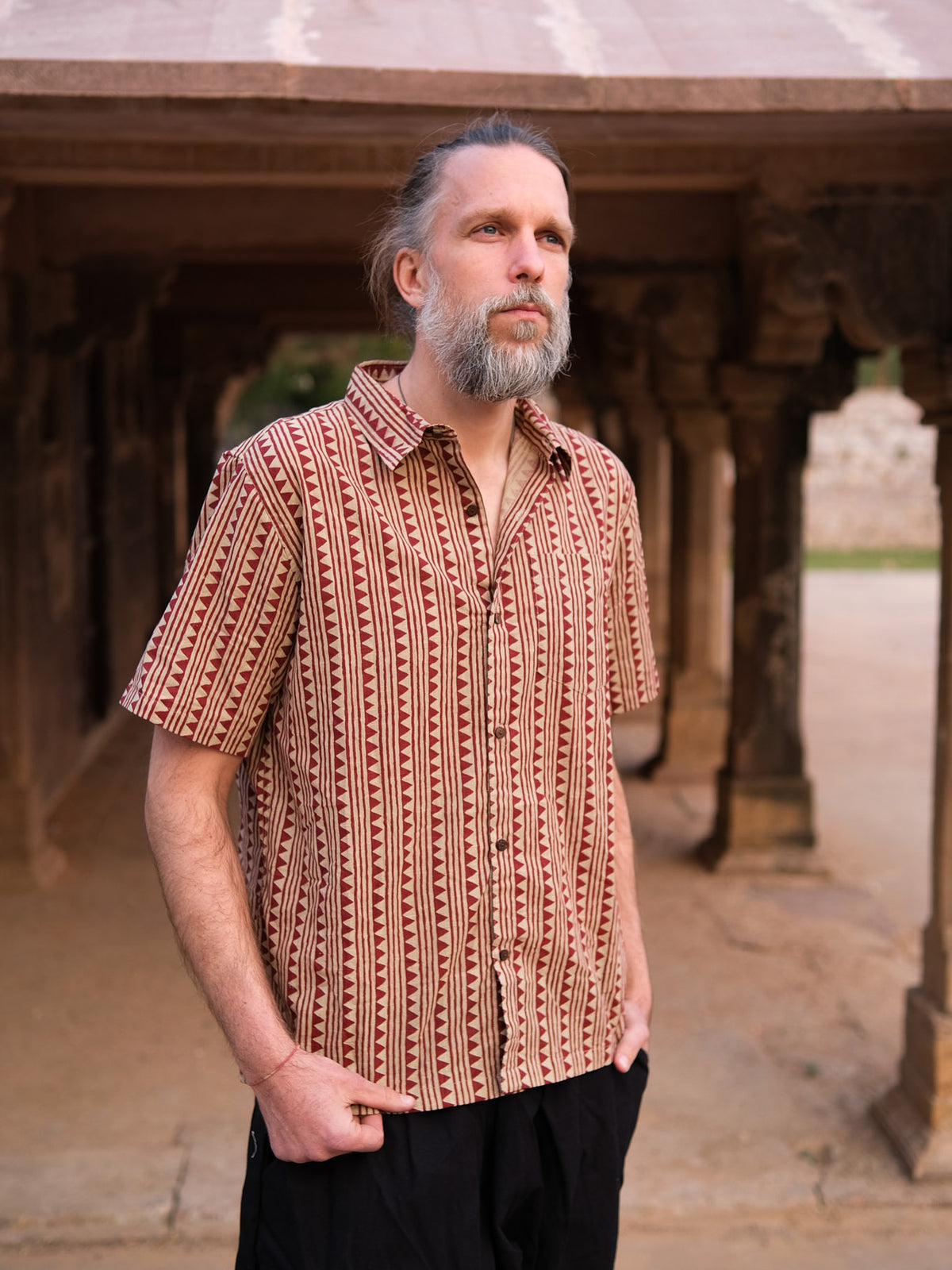 A man with gray hair and a beard stands outdoors beneath a stone structure, wearing the Tribal Triangle shirt—a unisex top that showcases a vibrant Rajasthan print—paired with black pants. He has his hands in his pockets and gazes into the distance.