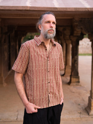A man with gray hair and a beard stands outdoors beneath a stone structure, wearing the Tribal Triangle shirt—a unisex top that showcases a vibrant Rajasthan print—paired with black pants. He has his hands in his pockets and gazes into the distance.
