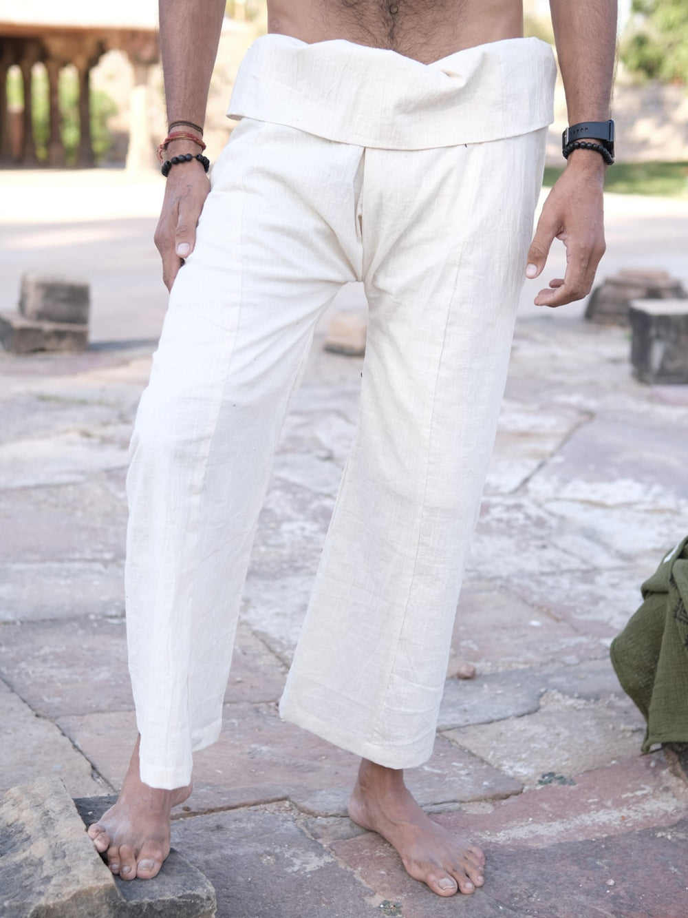 A person stands barefoot outdoors in loose-fitting, off-white Ren Thai Pants featuring a folded waistband, inspired by the traditional Thai silhouette. The high-quality Ren Thai Pants are set against a stone ground and blurred stone blocks in the background.