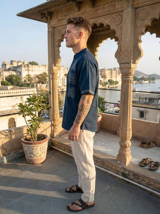 A man wearing the Midnight Nomad kurta, featuring handblock prints in blue, paired with beige pants, stands on a balcony with ornate arches overlooking historic buildings and water on a sunny day, embodying contemporary nomadic fashion.