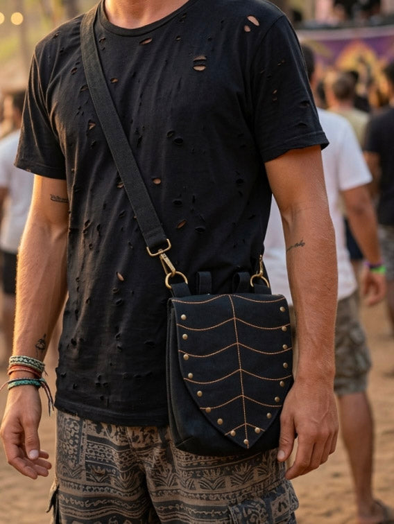 A person wearing a black, distressed t-shirt and patterned shorts stands outdoors, carrying the Luna Festival Bag in black—a utility sling bag featuring a leaf-flap design with gold studs that create a striking, shield-like effect.