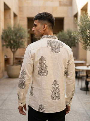 A man stands outdoors with his back to the camera, wearing the Ivory Crestwood Shirt. The shirt features a black heritage motif print on breathable natural fabric, perfectly suited to the relaxed setting of potted plants and empty tables in the background.