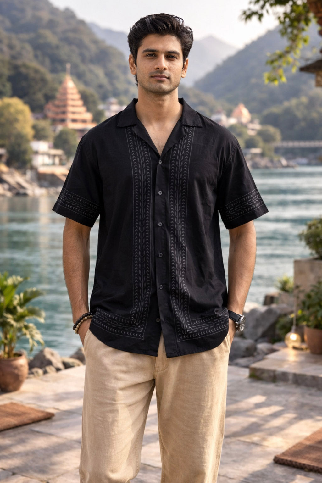 A man stands outdoors near a river, wearing the Zuri Shirt—a black hand block printed shirt—paired with beige pants. Lush green hills and temple-like buildings are visible in the background.