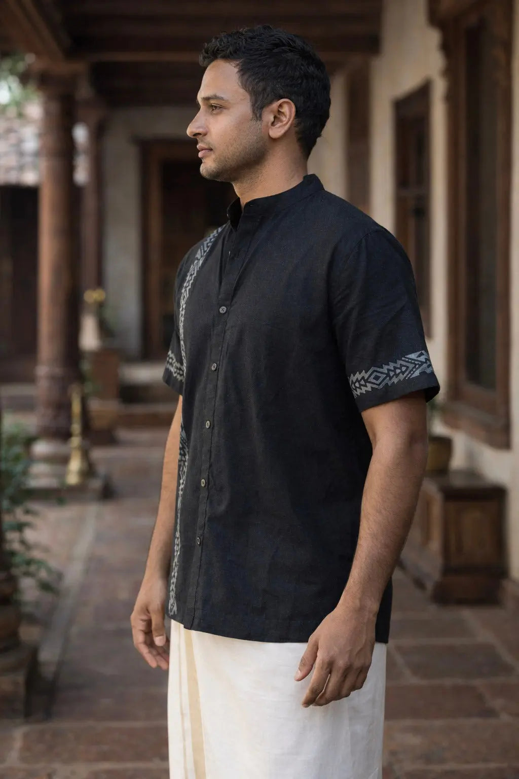 A man stands sideways in a corridor, wearing the black Heritage Shirt—crafted from breathable cotton with patterned detailing on one sleeve—and a white traditional garment around his waist.