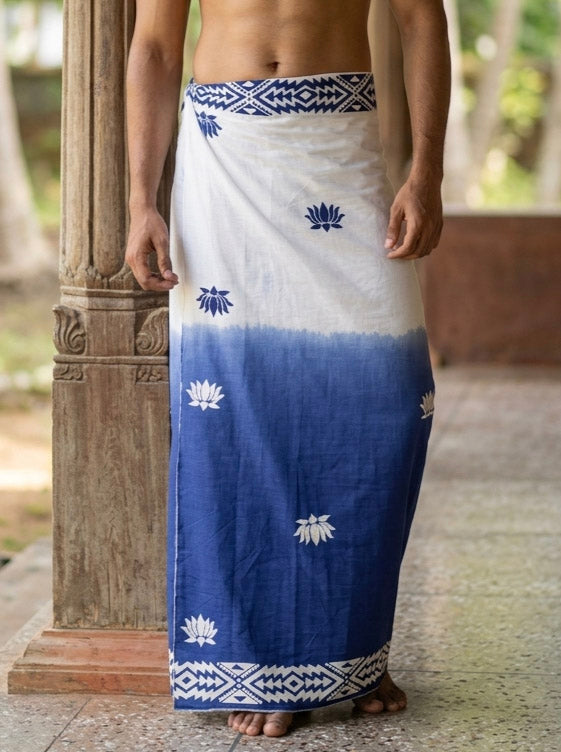 A person stands barefoot near a pillar, wearing the blue Sarovar Dhoti crafted from premium cotton and adorned with lotus flower patterns and a geometric border.