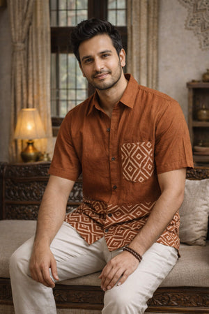 Wearing the Uday Shirt, a man in an earthy rust, hand block printed style pairs it with light pants as he sits on a beige sofa, smiling with a lamp and decorative items in the background.