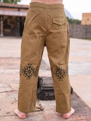 A barefoot person stands on stone, wearing sand yellow Tribal pants made from 100% cotton. The pants feature black hand blockprinted geometric patterns on the back and a right flap pocket. Outdoor walls and buildings are in the background.