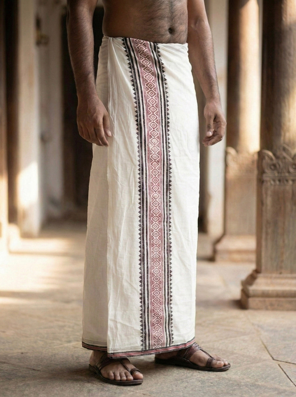 A man wearing the Anjana Dhoti—a handwoven cotton garment featuring a patterned border—stands barefoot in a corridor with stone pillars, evoking the timeless elegance of temple wear.