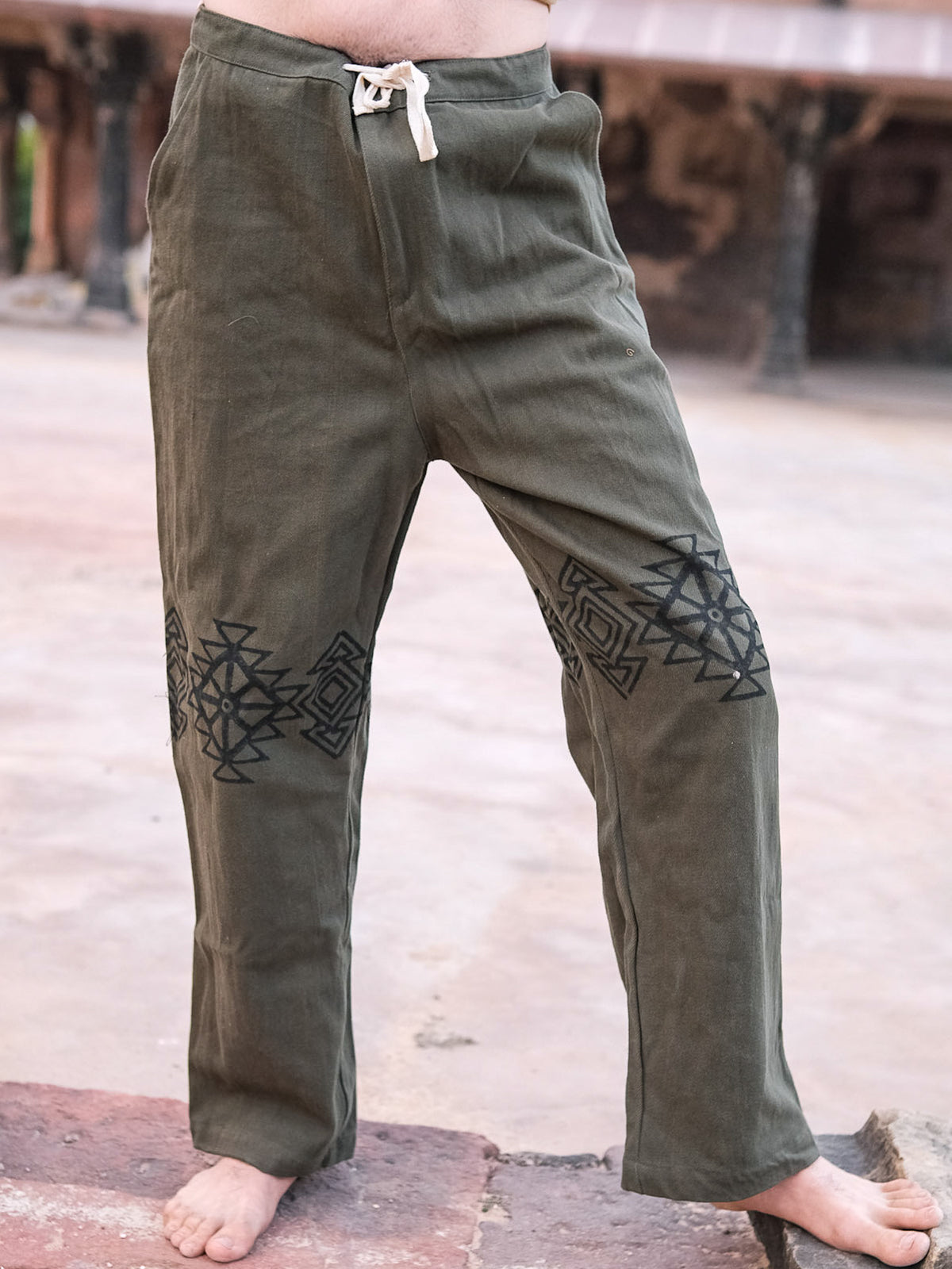 A person stands barefoot on stone, showing only their lower torso and legs dressed in olive green Tribal Pants—loose-fitting, 100% cotton with black geometric hand blockprints on the thighs.