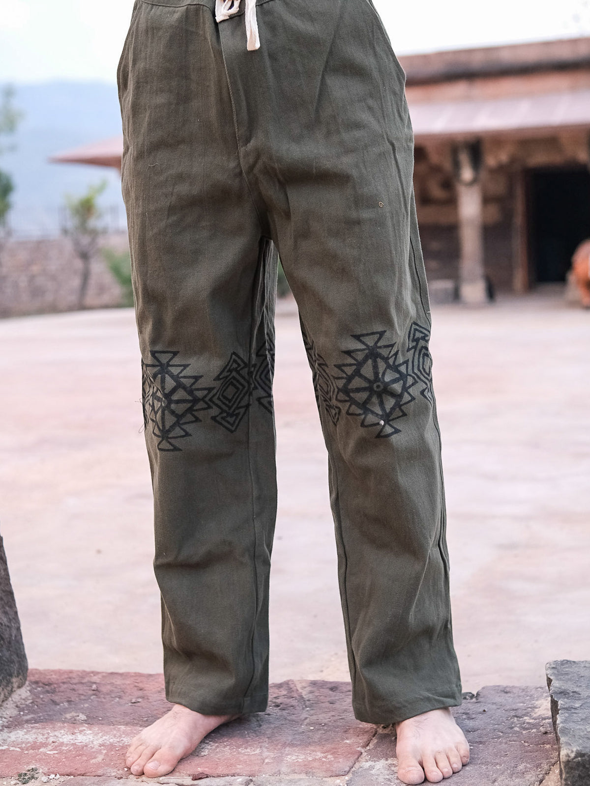 A person stands barefoot outdoors in green Tribal pants with black geometric embroidery at the knees and a drawstring waist, against a backdrop of paving stones and a rustic building.