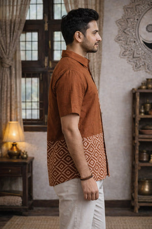A man stands in profile indoors, dressed in the Uday Shirt—an earthy rust hand block printed shirt featuring a geometric block print pattern at the bottom—paired with light pants. The room includes a window, shelves, a lamp, and decorative curtains.
