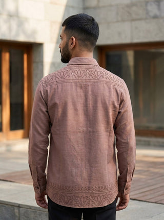 A man with short dark hair and a trimmed beard stands outdoors facing away, wearing the Madera shirt—a light brown, long-sleeve cotton piece featuring embroidered patterns on the back and sleeves. Crafted from breathable fabric, the Madera shirt is perfect for warm days. Stone walls and large windows are visible in the background.