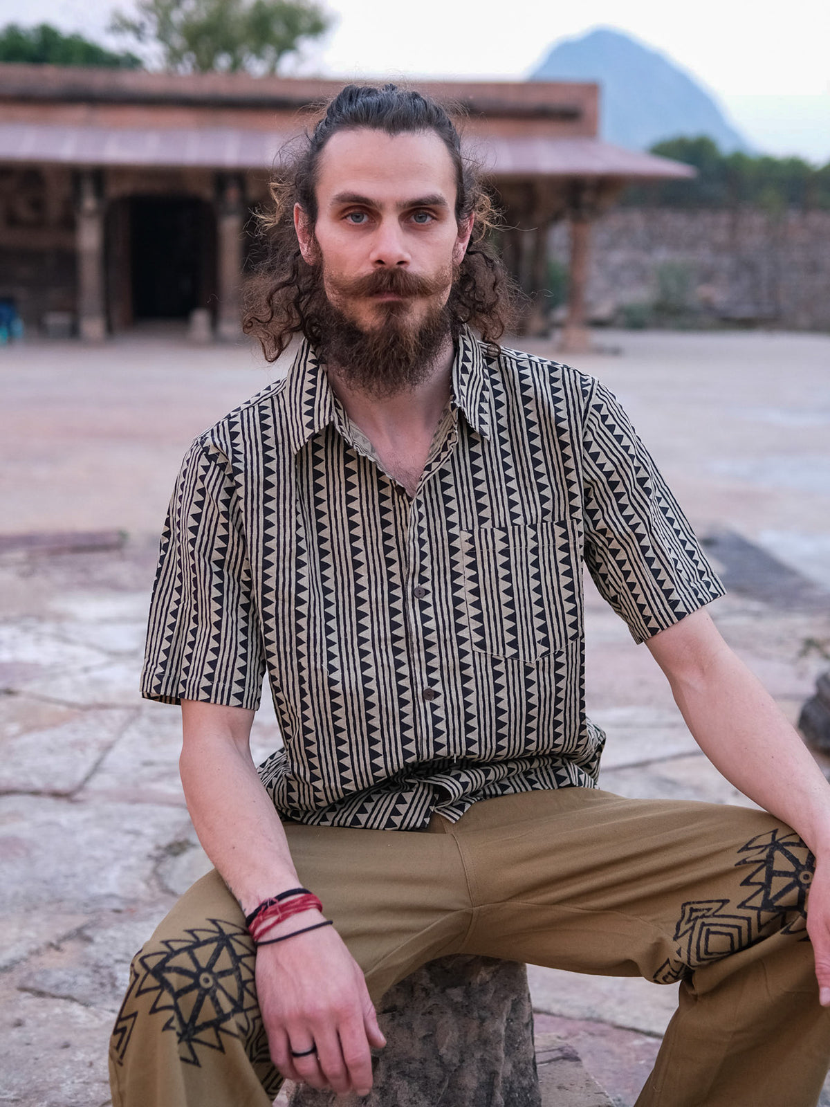 A man with curly hair and a beard sits outdoors on a stone, wearing the Tribal Triangle shirt—a stylish unisex short-sleeve top with Rajasthan-inspired patterns—paired with tan pants featuring black designs. A building and trees are visible in the background.