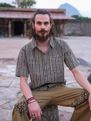 A man with curly hair and a beard sits outdoors on a stone, wearing the Tribal Triangle shirt—a stylish unisex short-sleeve top with Rajasthan-inspired patterns—paired with tan pants featuring black designs. A building and trees are visible in the background.