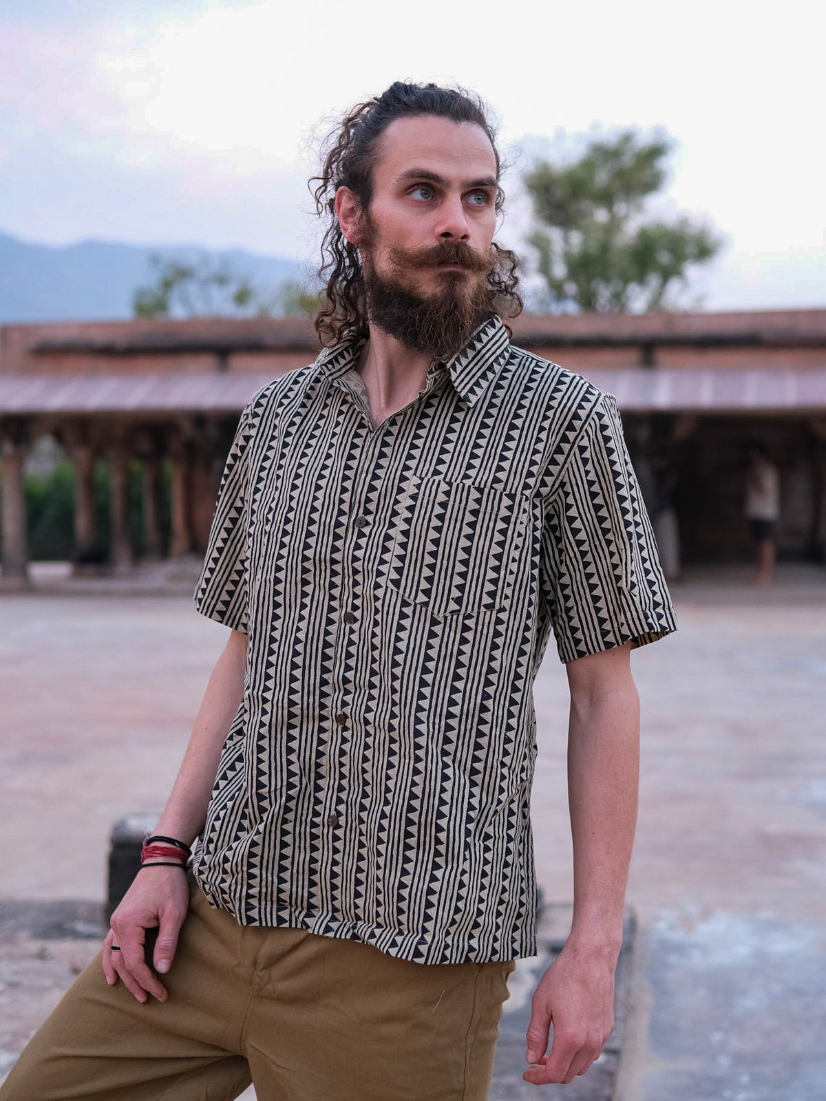 A man with curly hair and a beard is wearing the Tribal Triangle shirt with a Rajasthan-inspired print, paired with khaki pants, as he stands outdoors among trees and rustic buildings.